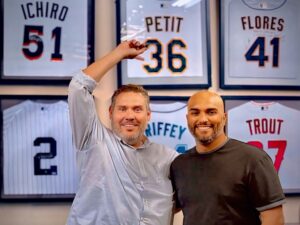 Eliam and Yusmeiro standing along a wall of jerseys with Eliam pointing at the Petit 36 jersey